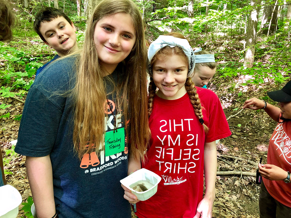 Three students smile as they hold up a container they're using to collect organisms from the creek during a creek study.
