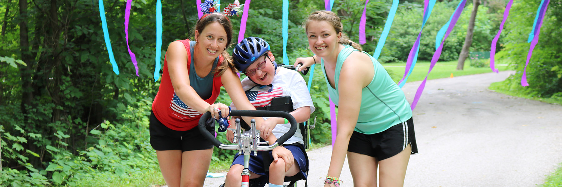 A smiling camper is helped by two counselors on an adapted bicycle.